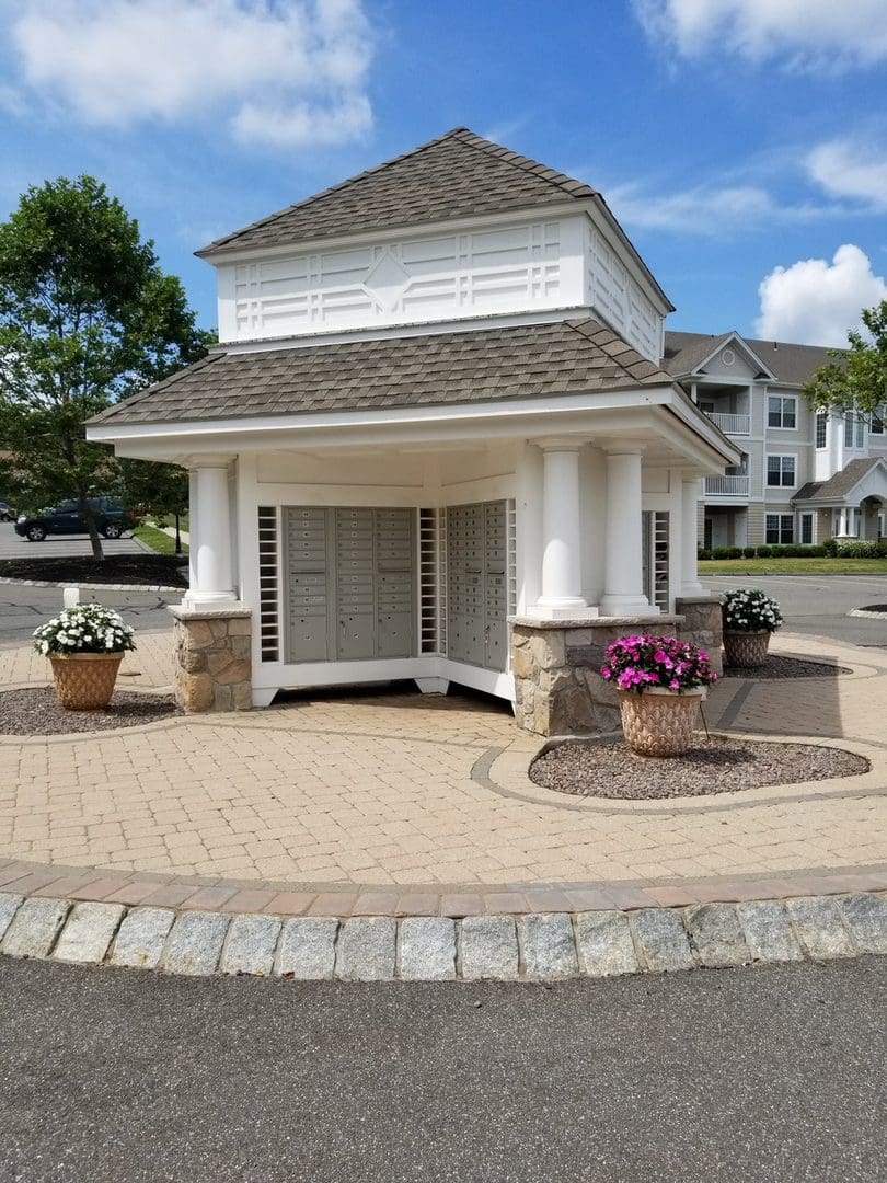A small, elegant pavilion with a white cupola and stone foundations flanked by potted plants, on a sunny day with residential buildings in the background.
